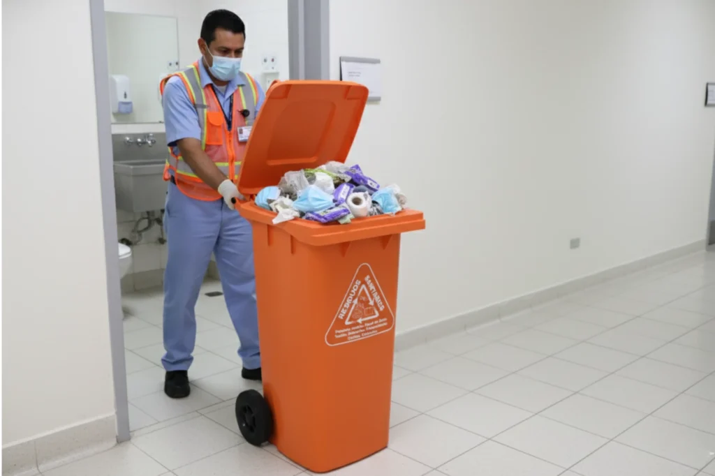 Trabajador empujando un contenedor naranja lleno de residuos sanitarios en un hospital para su correcta disposición.
